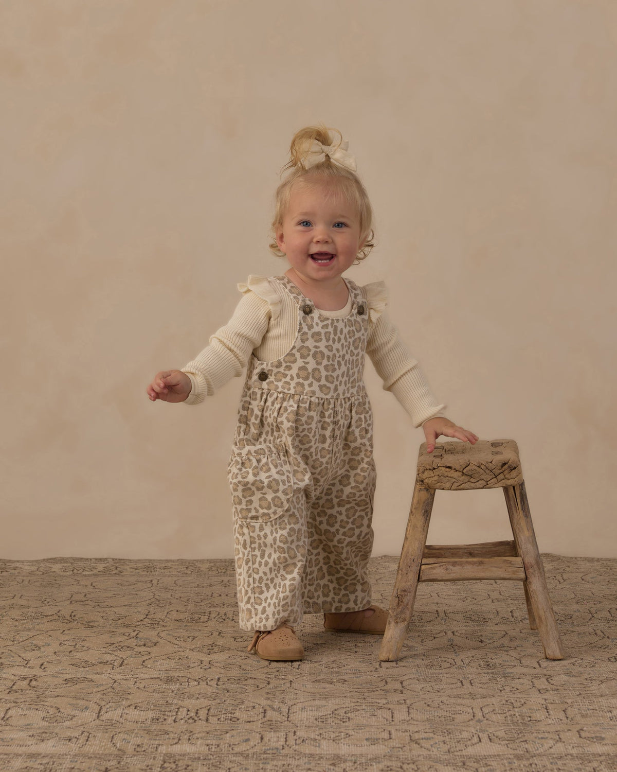 toddler girl wearing beige and brown leopard-print overall with a cream long-sleeve shirt stands on a textured beige floor next to a small wooden stool.