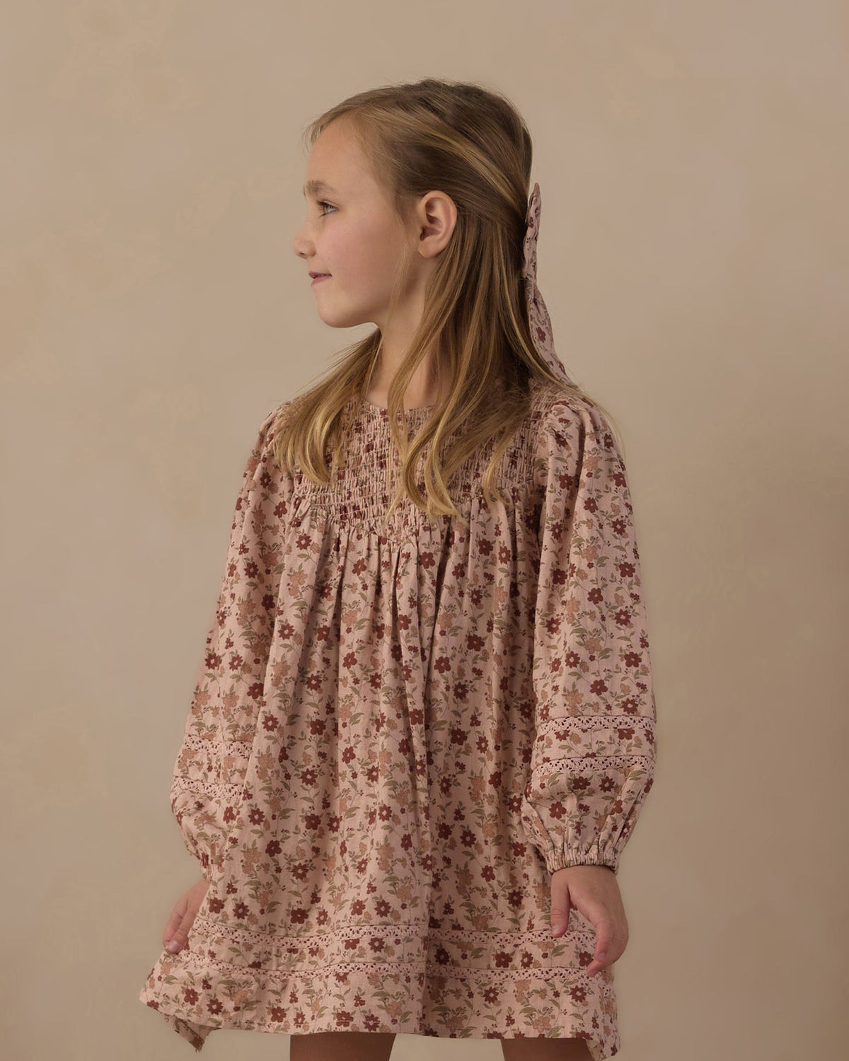A young girl with blonde hair tied back is wearing a long-sleeved, smocked babydoll dress with a dusty rose and brown floral pattern, photographed against a neutral beige background.