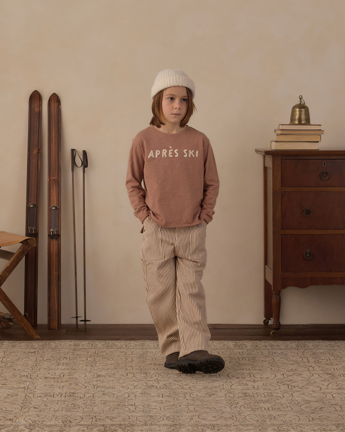 A child stands in a styled indoor room wearing a dusty rose long-sleeve sweatshirt with 'APRÈS SKI' printed in white across the chest, paired with beige corduroy pants and a white knit beanie. Ski boots and skis lean against the wall on the left, and a wooden cabinet with books and a small brass bell sits on the right.