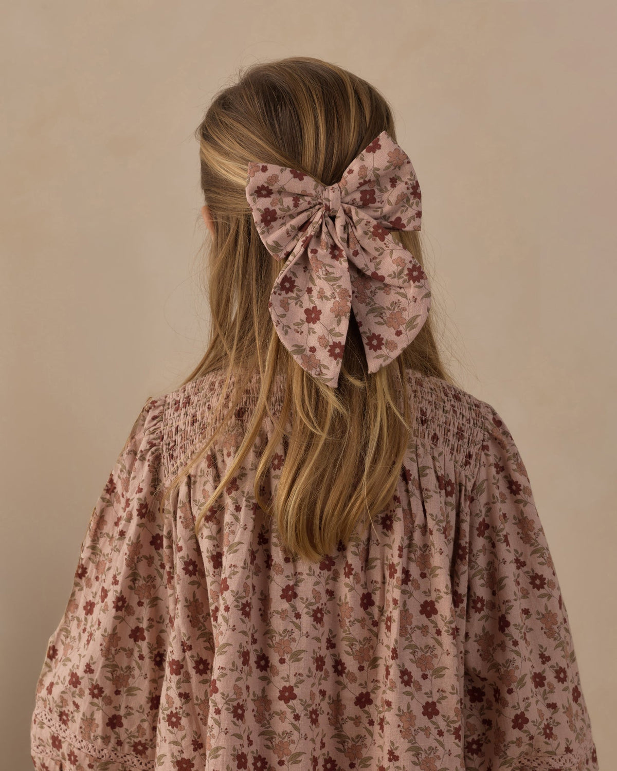 A blonde girl viewed from behind, wearing a large dusty pink floral hair bow accessory in her hair, paired with a matching smocked blouse.