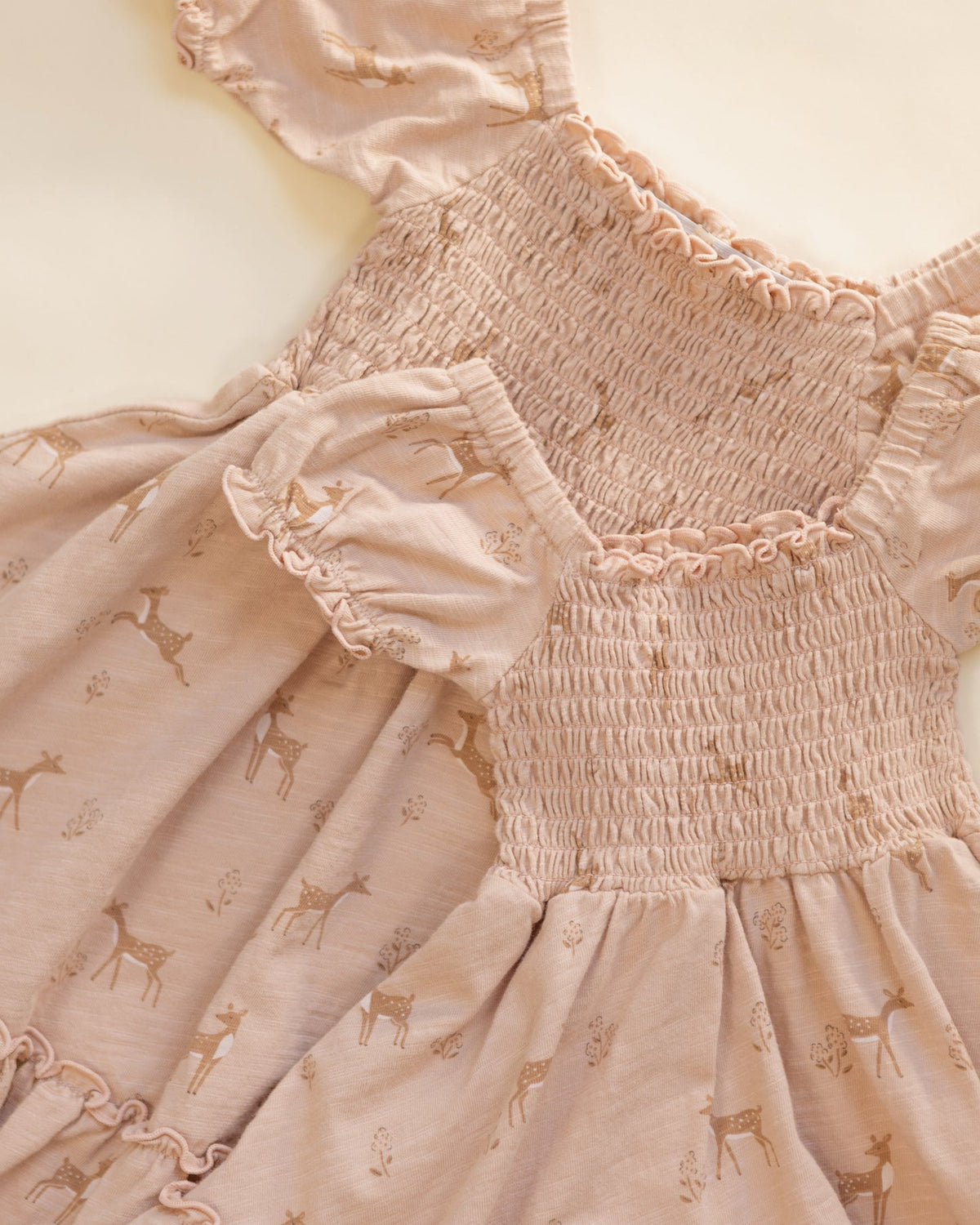 A close-up of a baby dress in a dusty pink color, featuring a smocked bodice, ruffled trim, and a printed pattern of small deer and floral motifs.