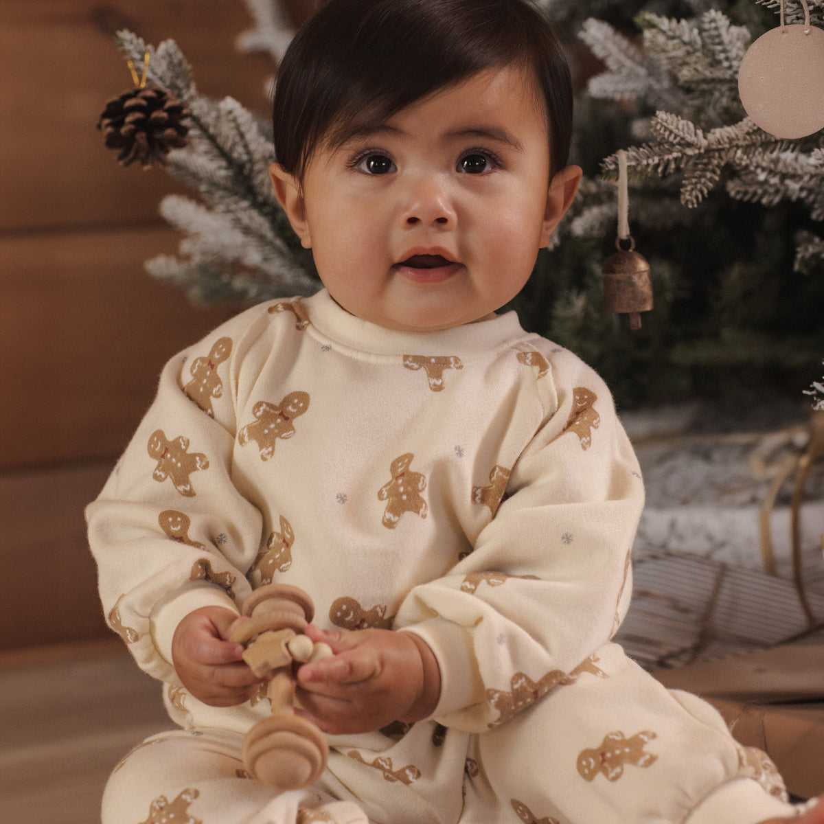 A baby sitting in front of a decorated Christmas tree, wearing a cream-colored long-sleeve pajama set with brown gingerbread cookie prints.