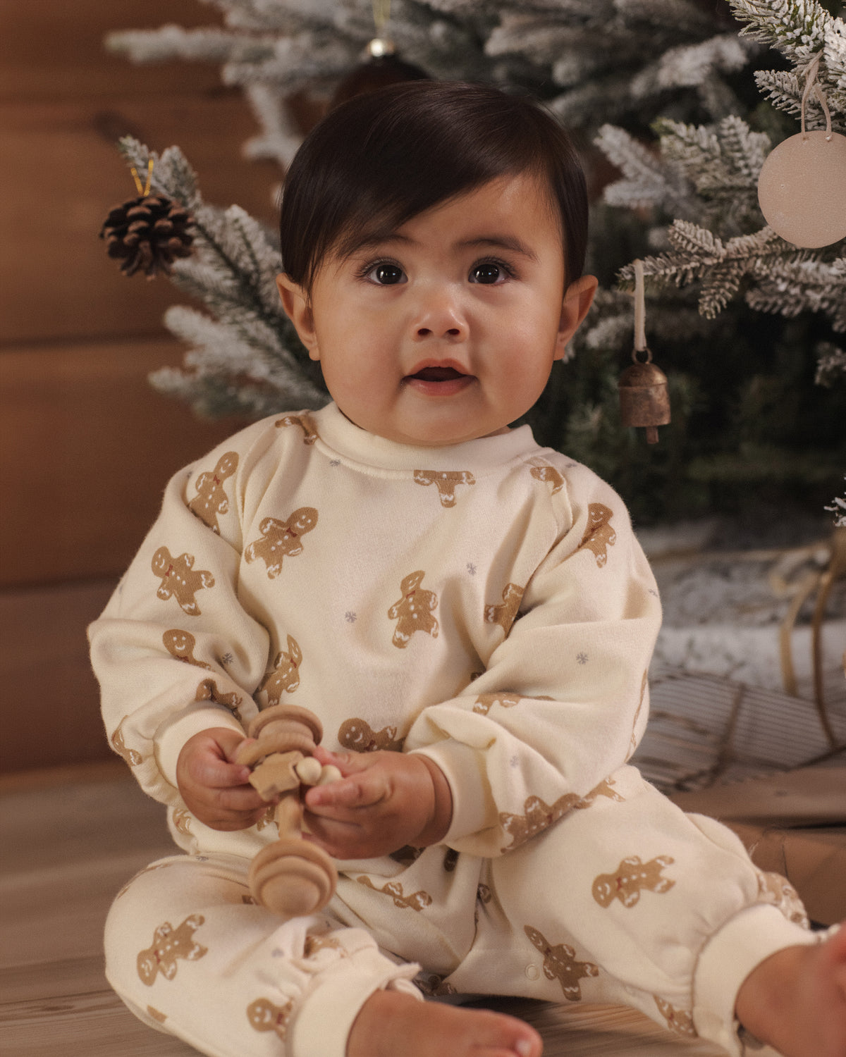 A baby sitting in front of a decorated Christmas tree, wearing a cream-colored long-sleeve pajama set with brown gingerbread cookie prints.