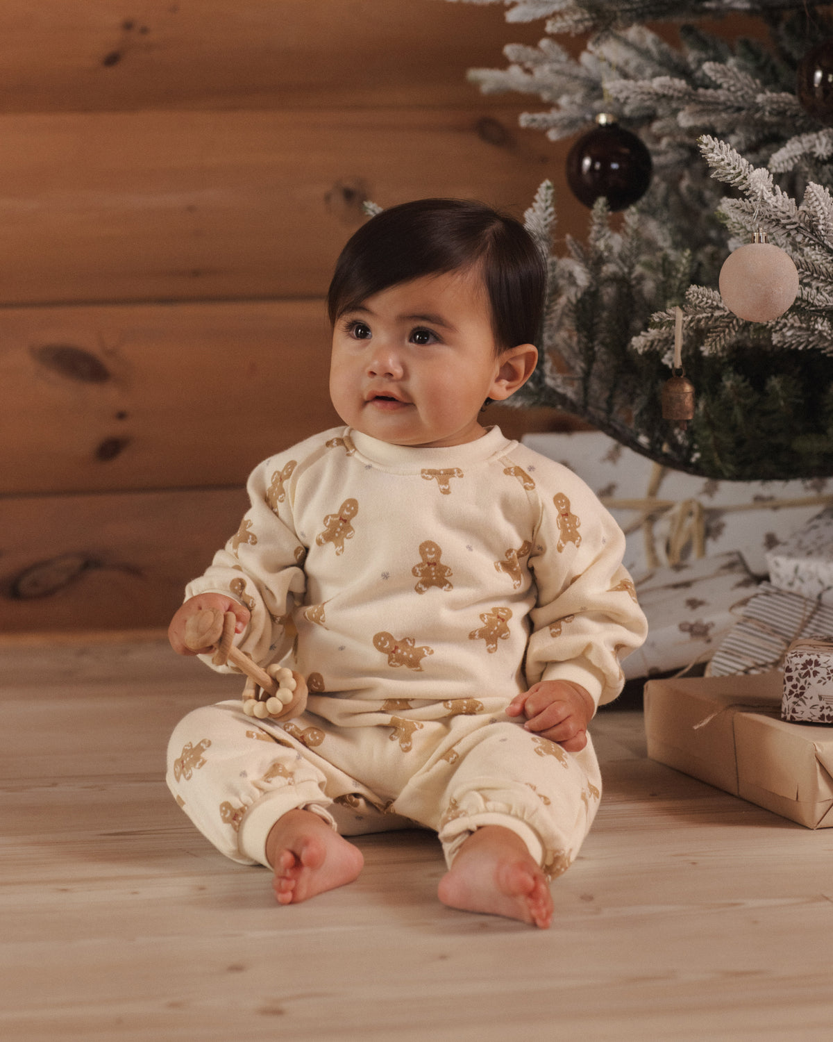 A baby sitting on a wooden floor in front of a Christmas tree, wearing a cream-colored one-piece pajama with a brown gingerbread man pattern.