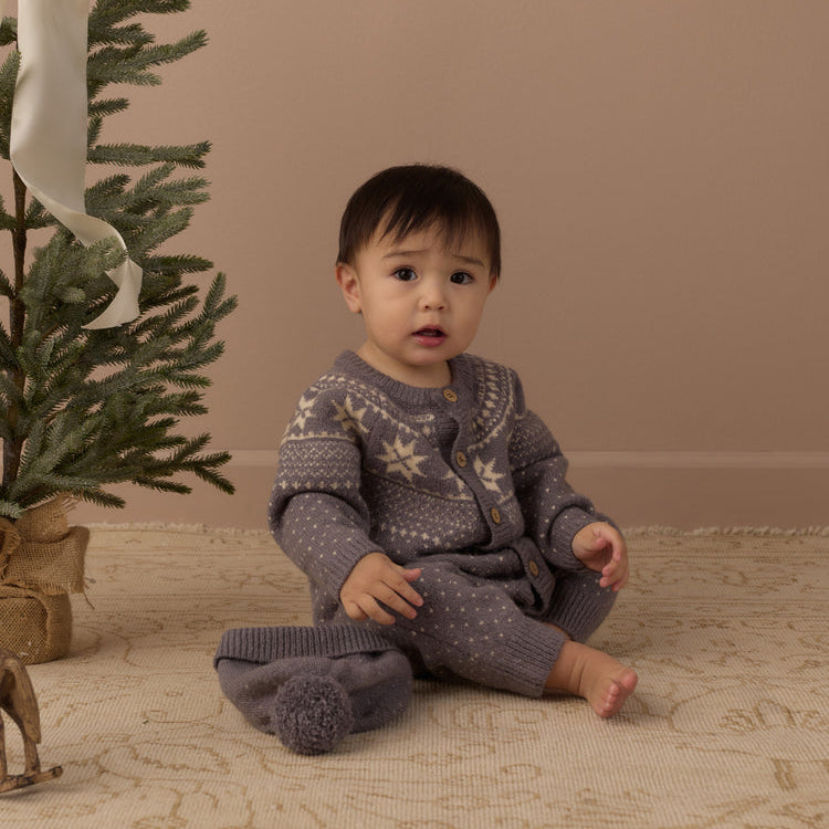 A baby sitting on a patterned rug next to a decorated Christmas tree, wearing a gray knit cardigan and matching knit pants with cream-colored snowflake patterns.