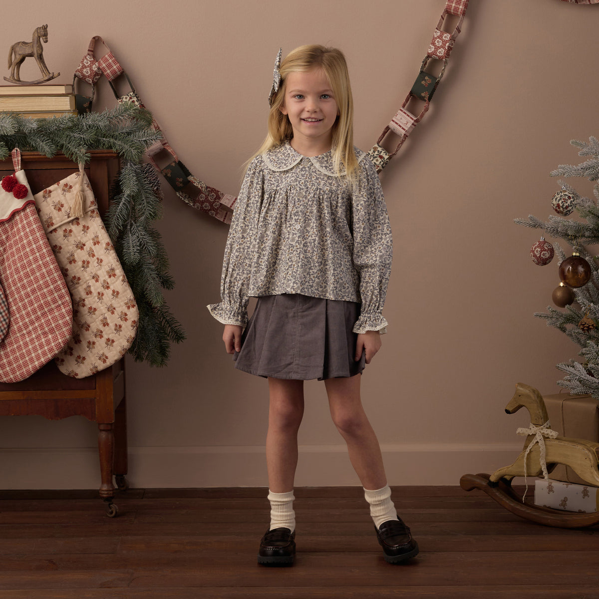 A young girl stands in a decorated indoor room wearing a long-sleeve patterned blouse with a Peter Pan collar and a dark gray skirt. The background features a warm beige wall with a garland, stockings, and a Christmas tree.