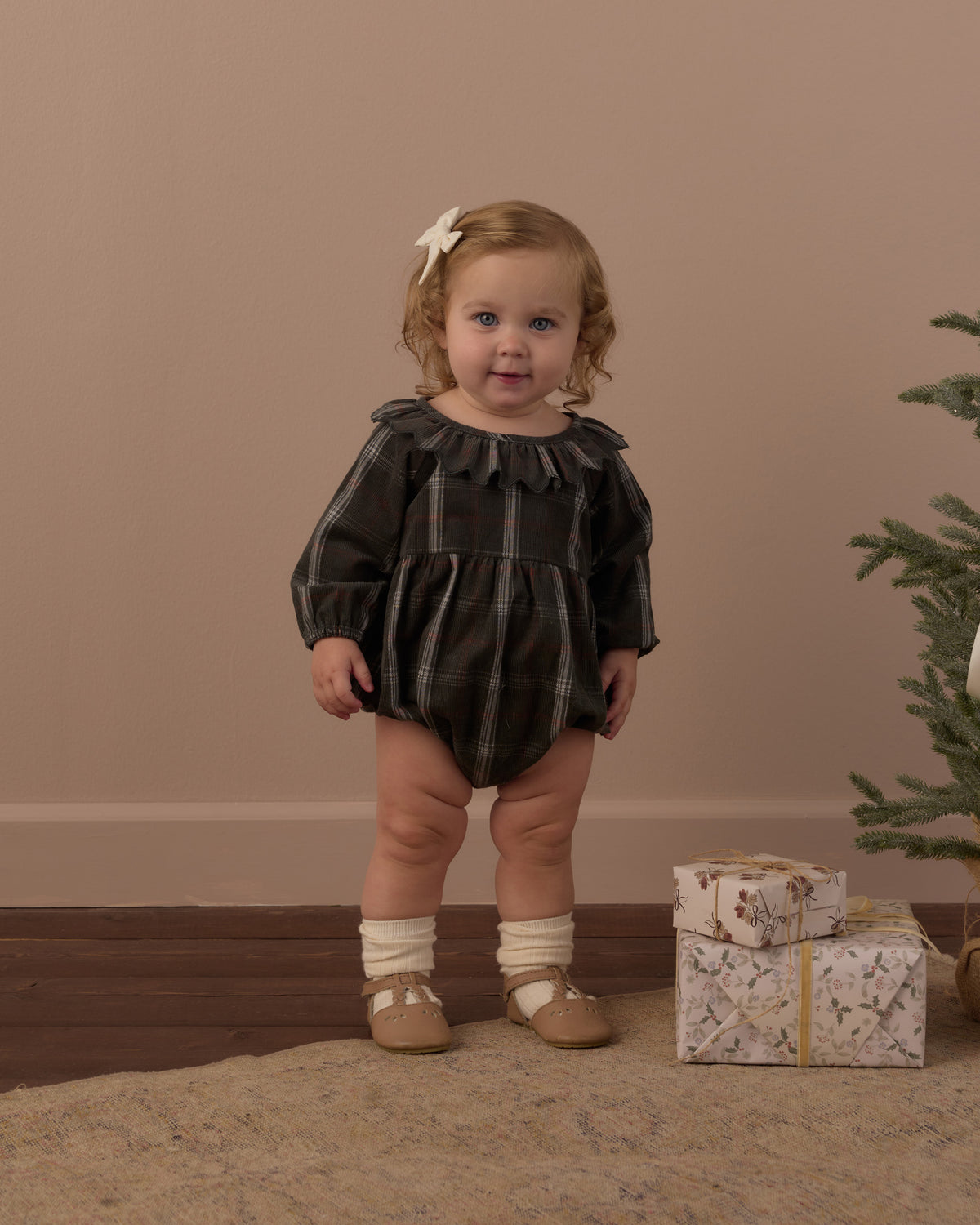 A toddler stands indoors wearing a dark plaid romper with long puff sleeves, cream socks, and tan shoes. The room has a warm beige wall, wood floor, a small Christmas tree, and wrapped gifts on the floor.