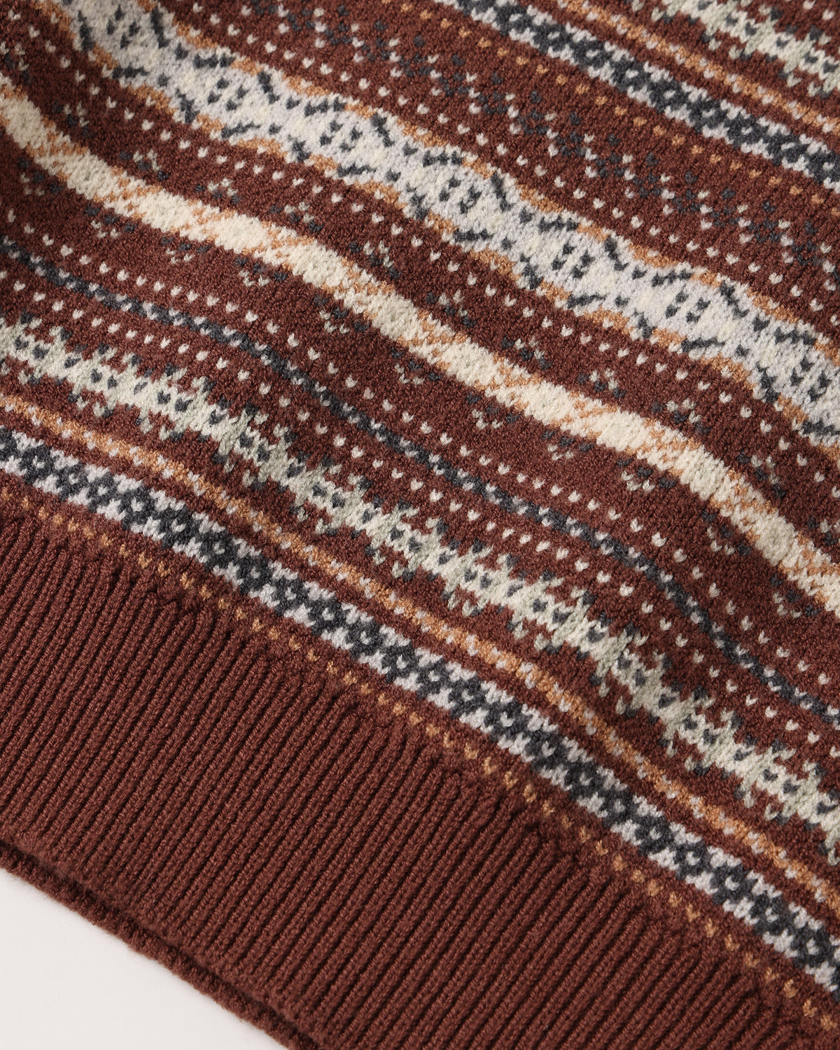 Close-up of a knit sweater corner showing a brown ribbed cuff and a Fair Isle style pattern with horizontal stripes in brown, cream, and dark gray.