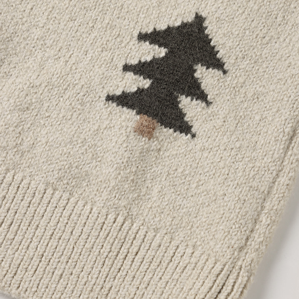 A close-up view of a cream-colored knit sweater featuring a dark green pine tree pattern with a brown trunk and ribbed texture at the hem.