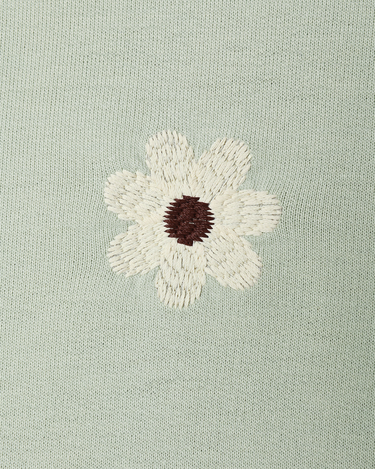 A close-up of a light sage green knit fabric featuring an embroidered daisy flower with cream petals and a dark brown center.