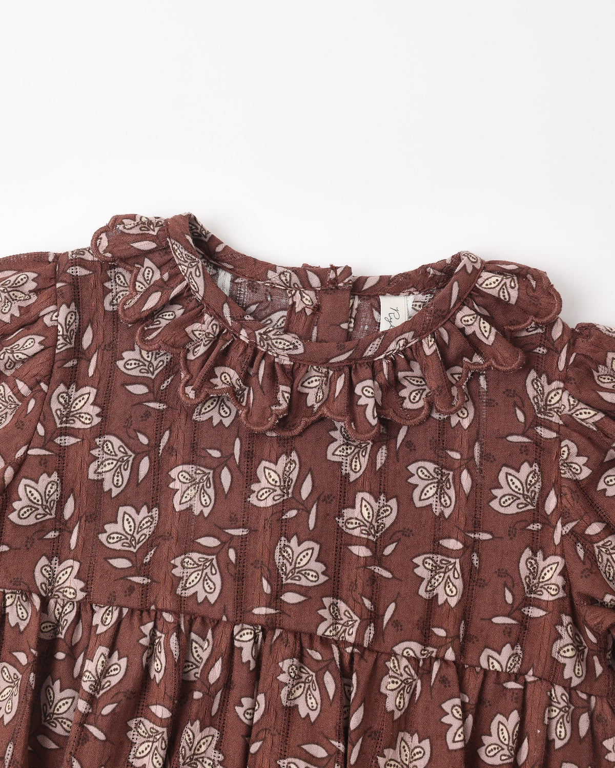 A close-up of a brown cotton dress with a white floral and leaf pattern, featuring a ruffled collar and tiered skirt.