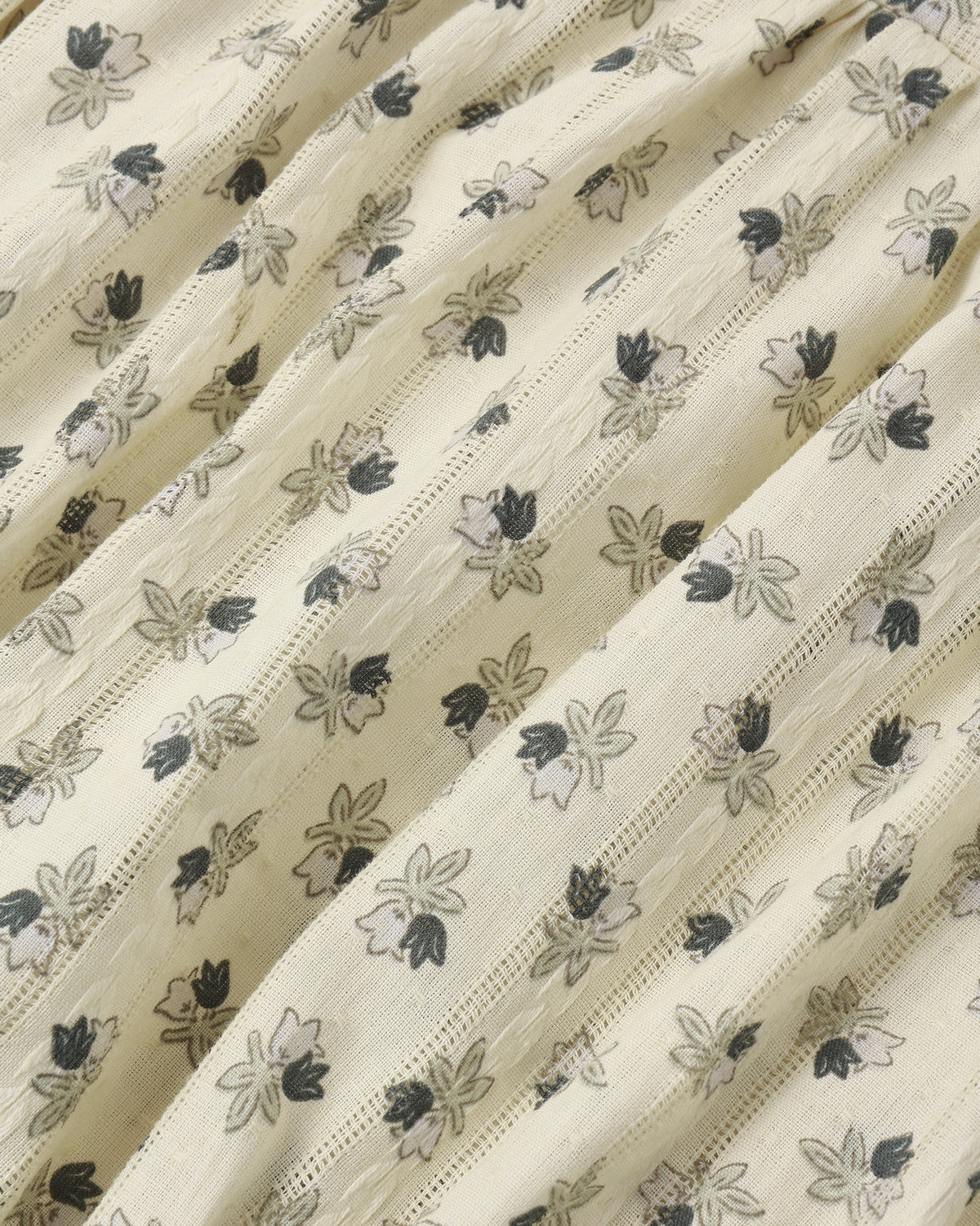 A close-up of a cream-colored fabric with a delicate, repeating floral block print in black and gray, featuring fine vertical stitched stripes.
