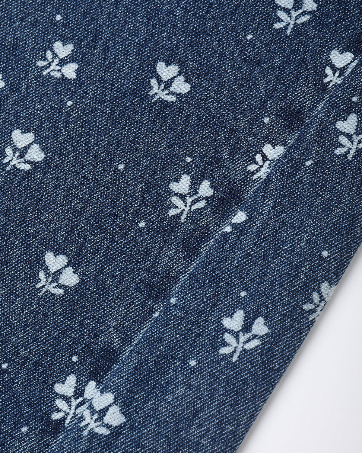 A close-up of dark blue denim fabric with a repeating white floral print and a diagonal seam.