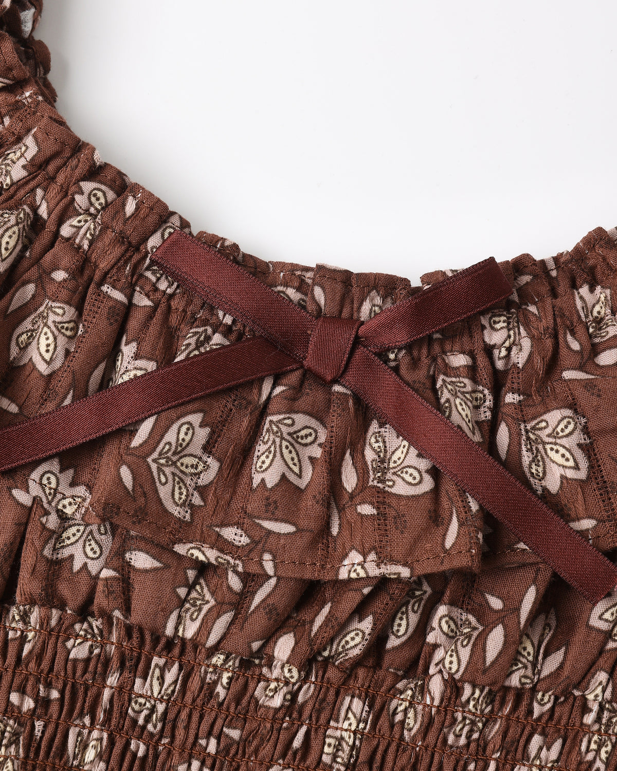 Close-up of a brown ruffled top with a beige floral print and a dark brown satin bow at the neckline, shown against a plain white background.
