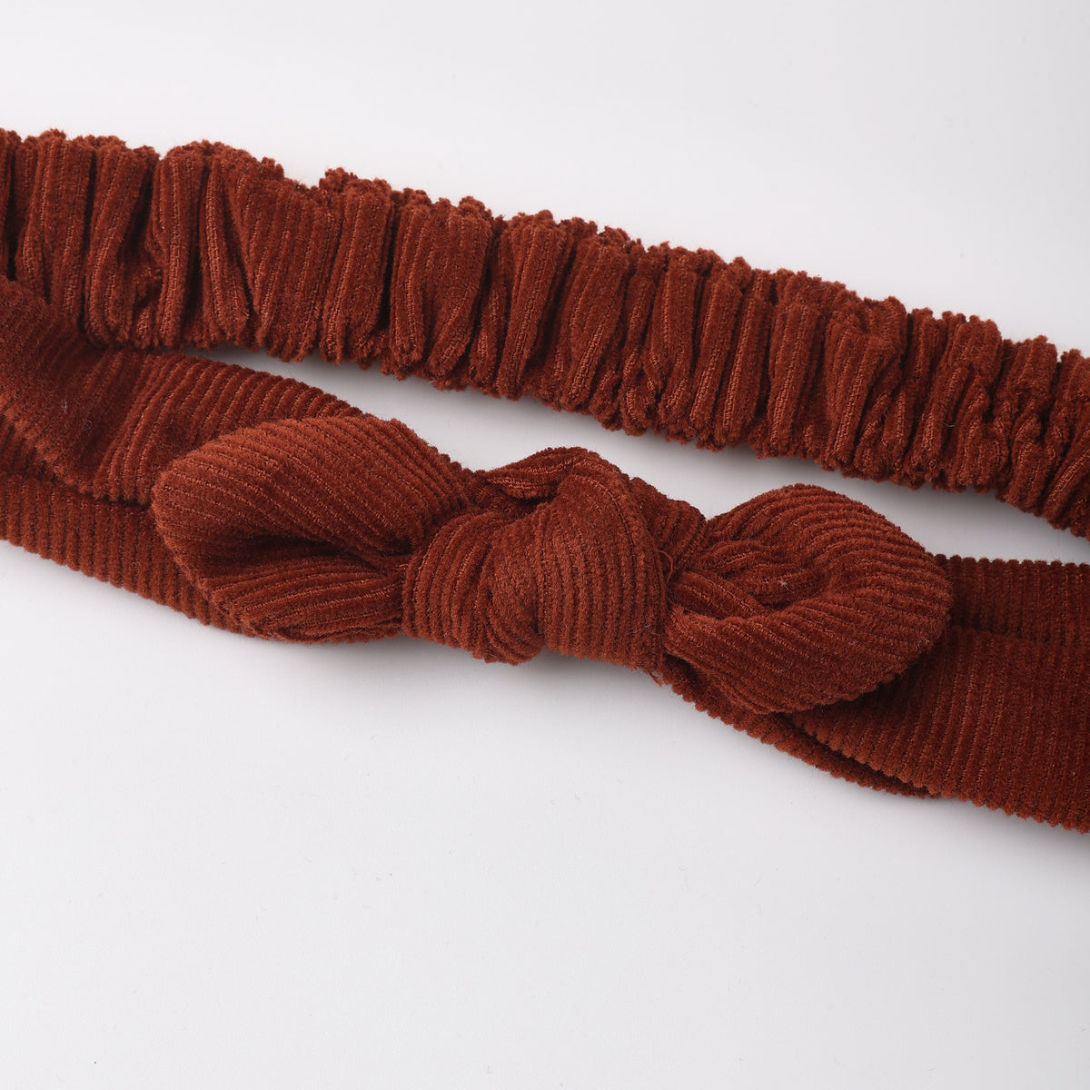 A rust-brown corduroy headband with a knotted bow detail and a matching elastic band, displayed on a clean white background.