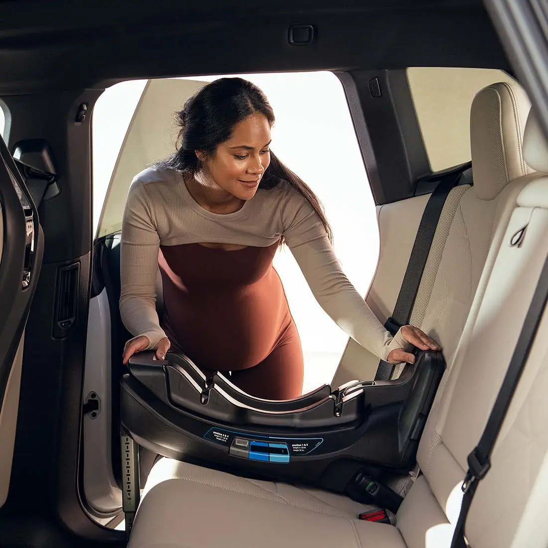 A pregnant woman leaning into the back seat of a car to install a MIXX Next + Pipa Aire RX Travel System by Nuna car seat base.