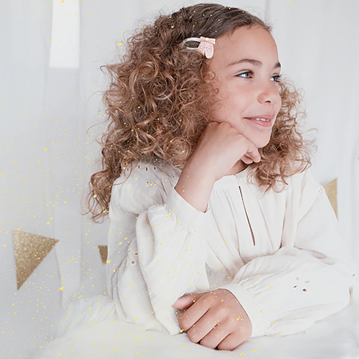 Young girl with curly hair wearing a white outfit with gold polka dots, sitting on a light-colored couch.