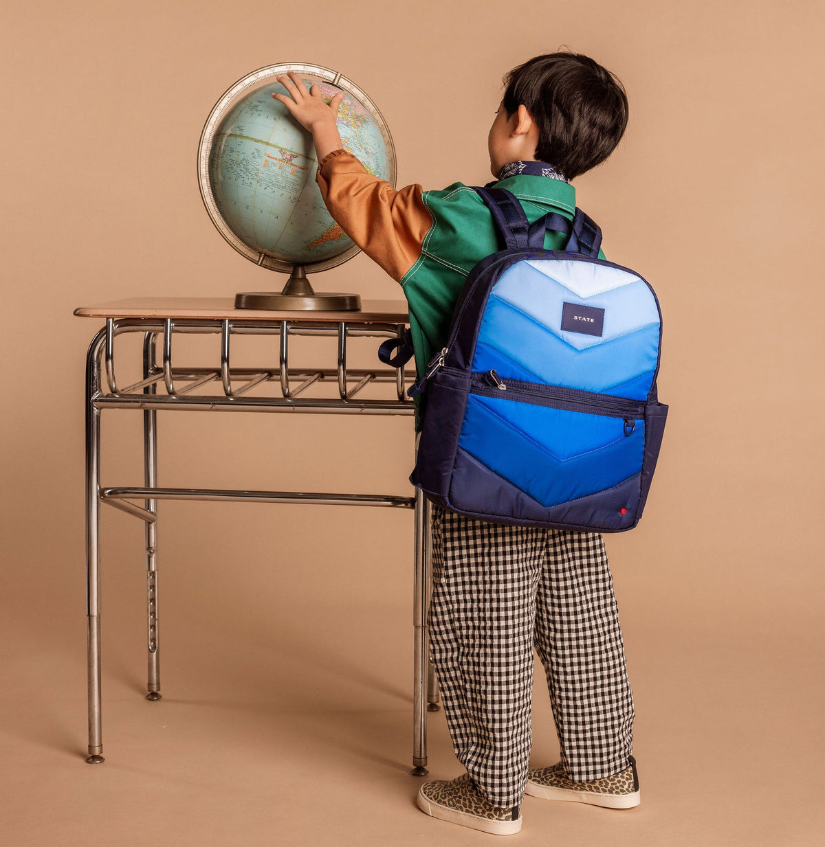 Child with a blue backpack looking at a globe on a desk against a beige background