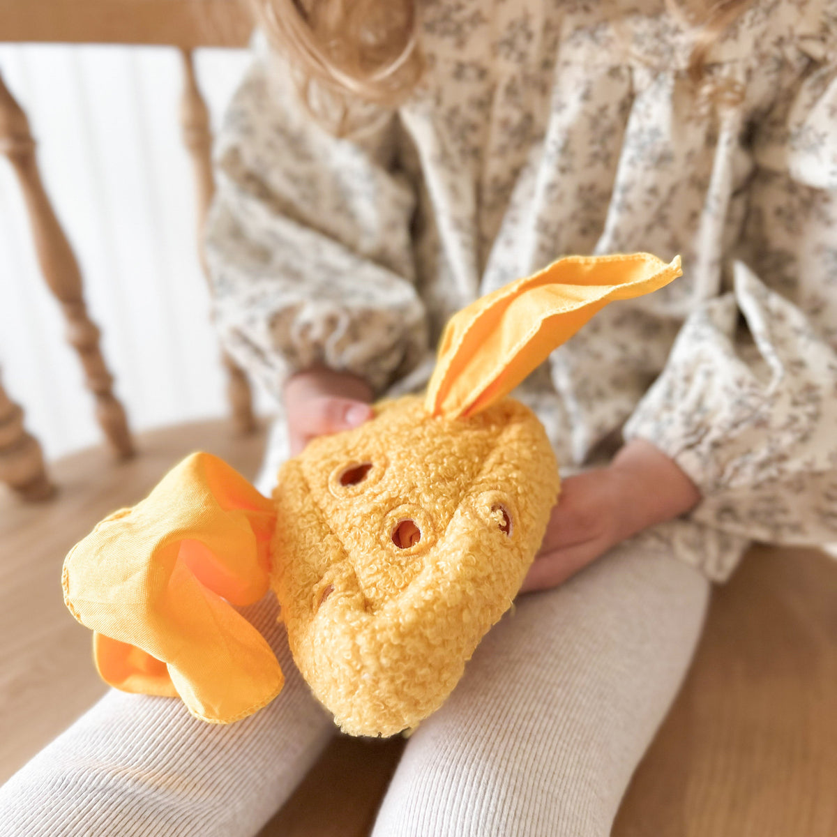 Child holding a plush carrot toy with a face on a wooden floor.