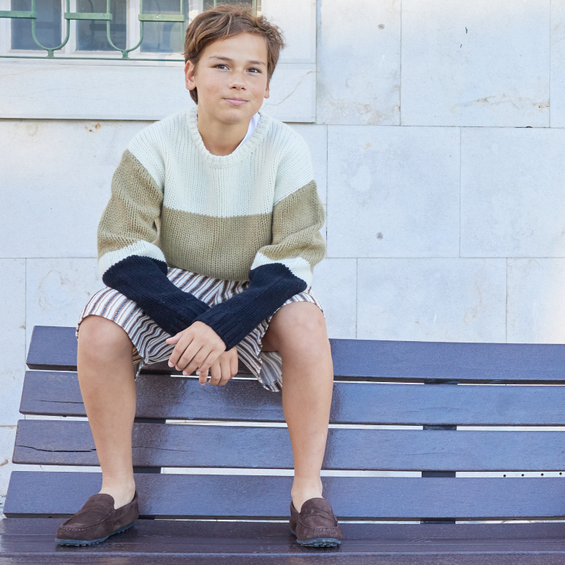 Young boy sitting on a blue bench wearing a striped sweater and shorts.