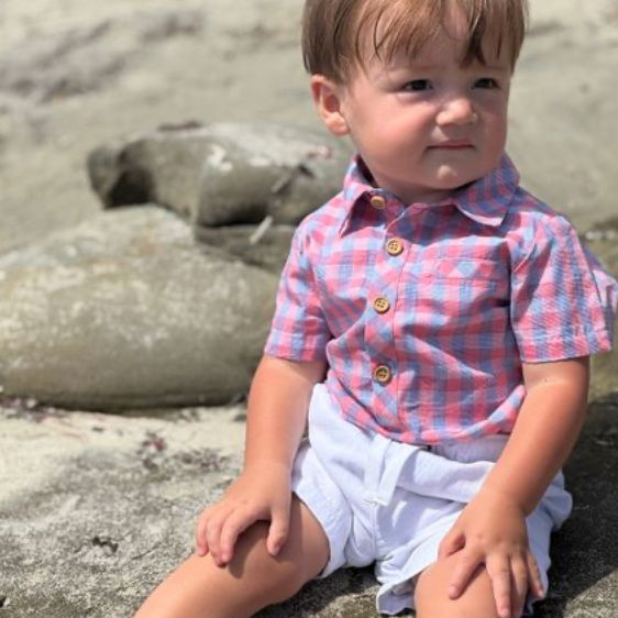 Child sitting on a rocky beach wearing a checkered shirt and white shorts.