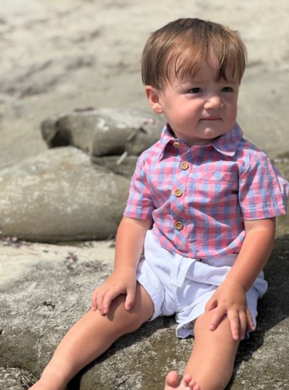 Child sitting on a rocky beach wearing a checkered shirt and white shorts.