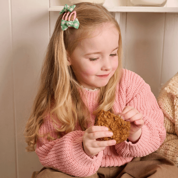 A young girl with long blonde hair wearing a pink knit sweater and a green hair clip, holding a piece of brown bread while sitting in a cozy indoor setting.