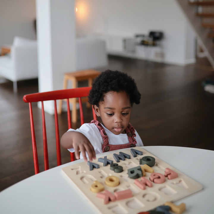 A toddler sitting at a table in a bright room, focused on placing a piece into a wooden alphabet puzzle.