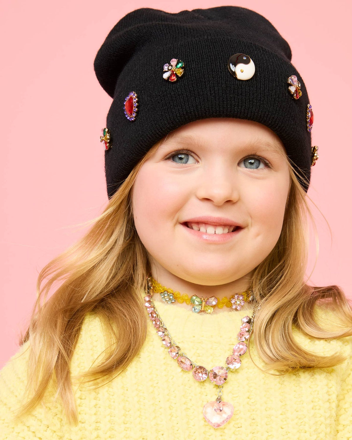 Child wearing a black beanie with colorful embellishments on a pink background