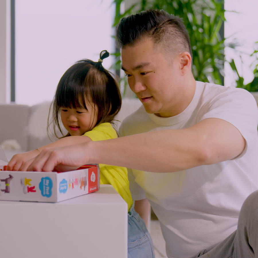 Man and young girl opening a box together in a bright room with plants in the background