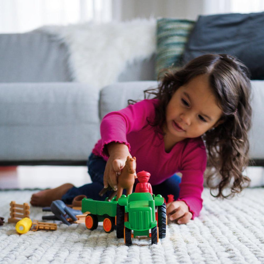 A young girl wearing a pink long-sleeve shirt smiling as she plays on the floor with the green Smartmax My First Farm Tractor set, featuring a brown toy horse and a red driver figure.