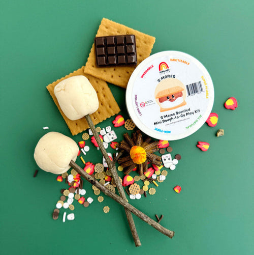 S'mores play set with marshmallows, graham crackers, chocolate bars, and a mini dough kit on a green background.