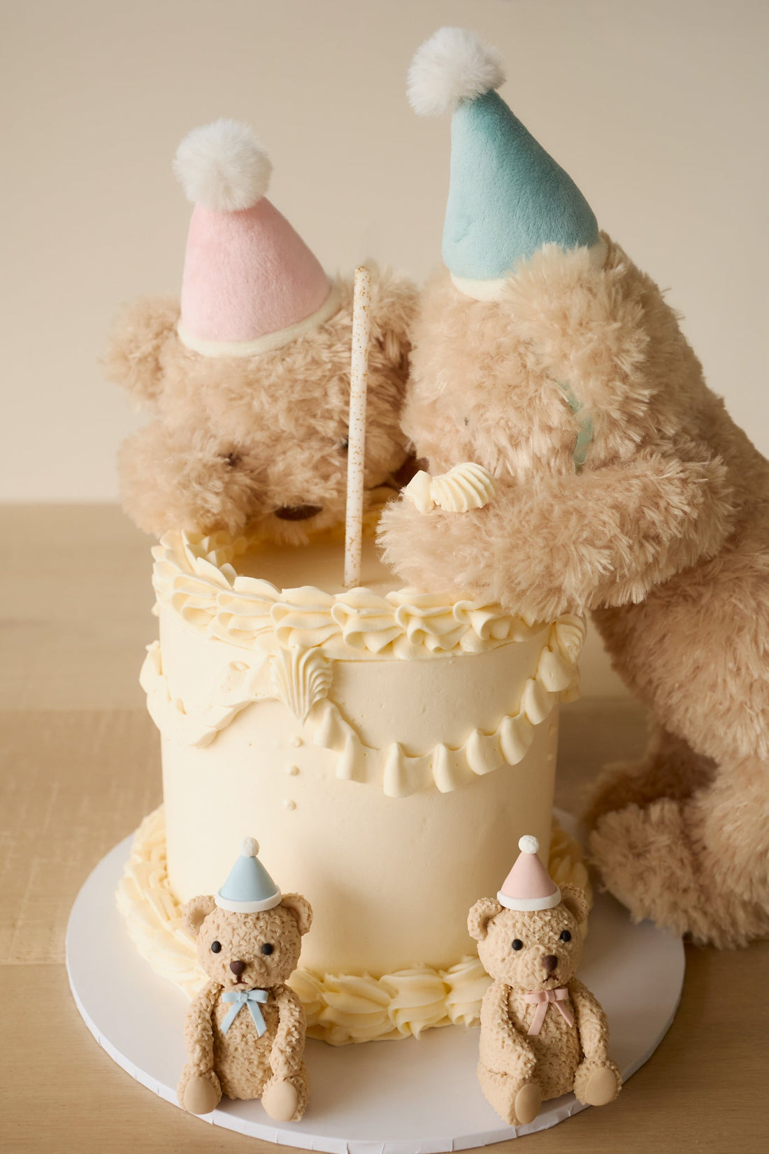 Two teddy bears with party hats around a cake on a wooden surface.