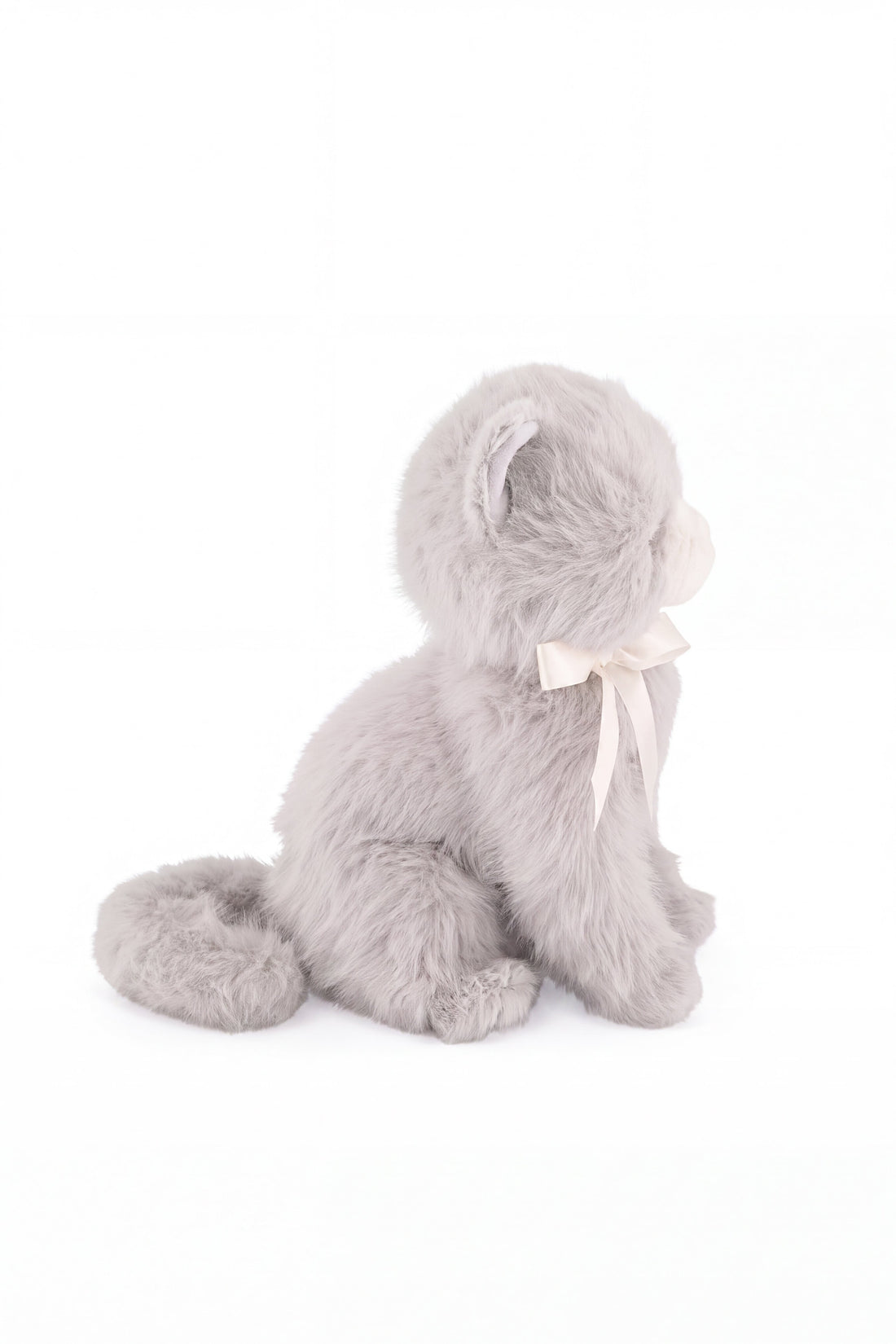 A soft grey plush kitten toy viewed in profile, showing its seated posture, curved tail, and a white satin ribbon bow tied around its neck.
