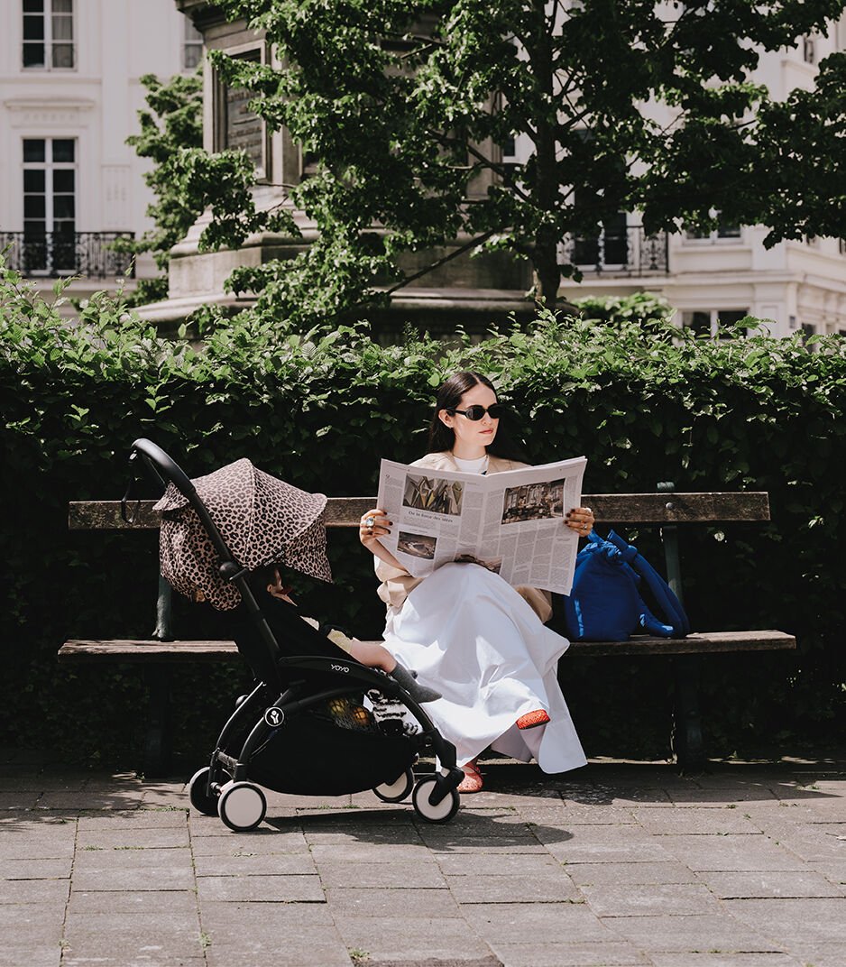Woman reading a newspaper on a bench with a stroller next to her