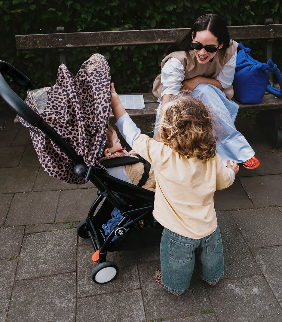 Woman sitting on a bench with a child next to a stroller, surrounded by greenery.
