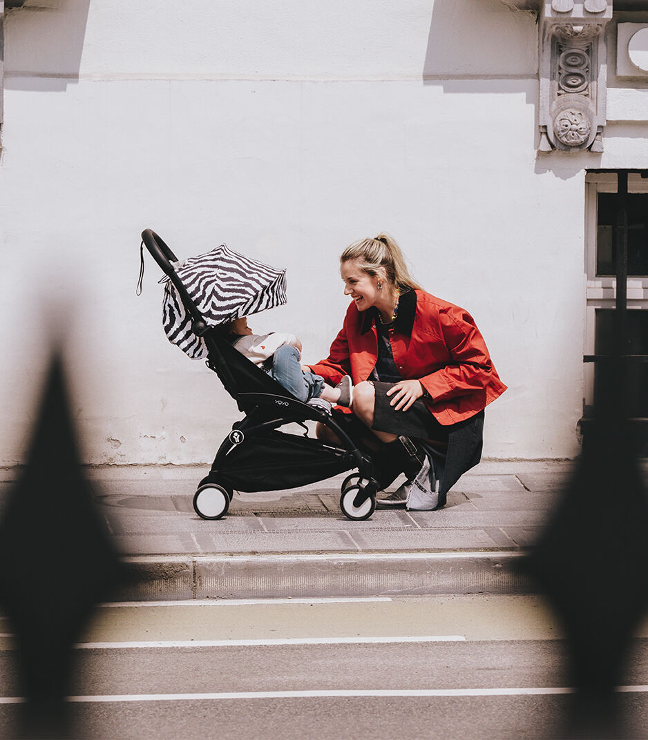 Woman in a red jacket sitting on steps with a child in a stroller, zebra-striped cover, against a white wall.