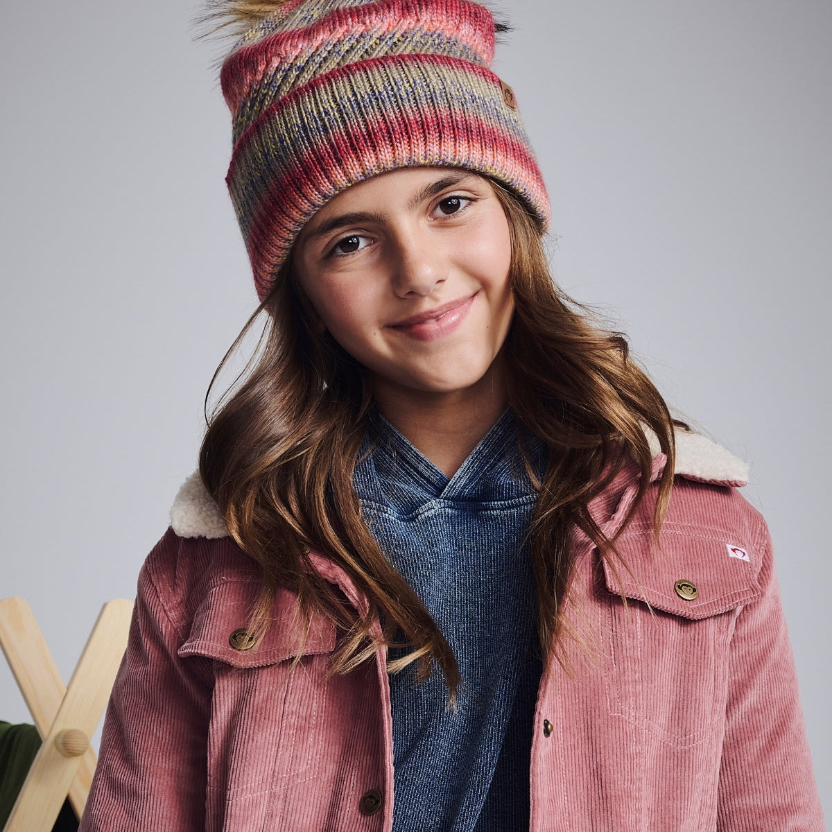 Young girl wearing a colorful knit hat and pink jacket against a gray background
