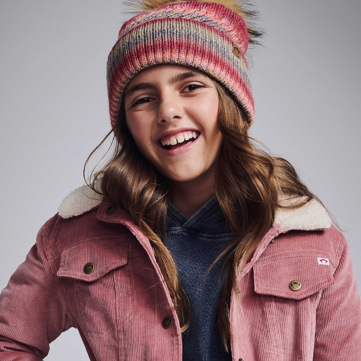 Young girl wearing a pink coat and colorful knit hat with pom-pom on a gray background