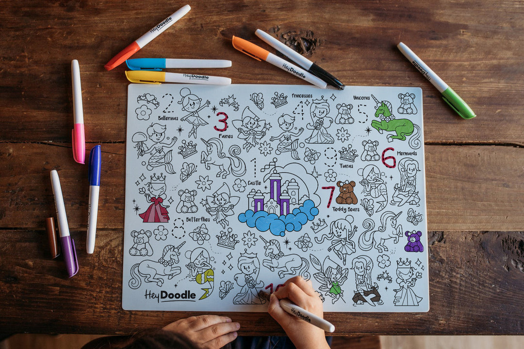 A child's coloring page with black line art illustrations and numbers, being drawn on by a hand using a marker, placed on a rustic wooden table with several colored markers around it.