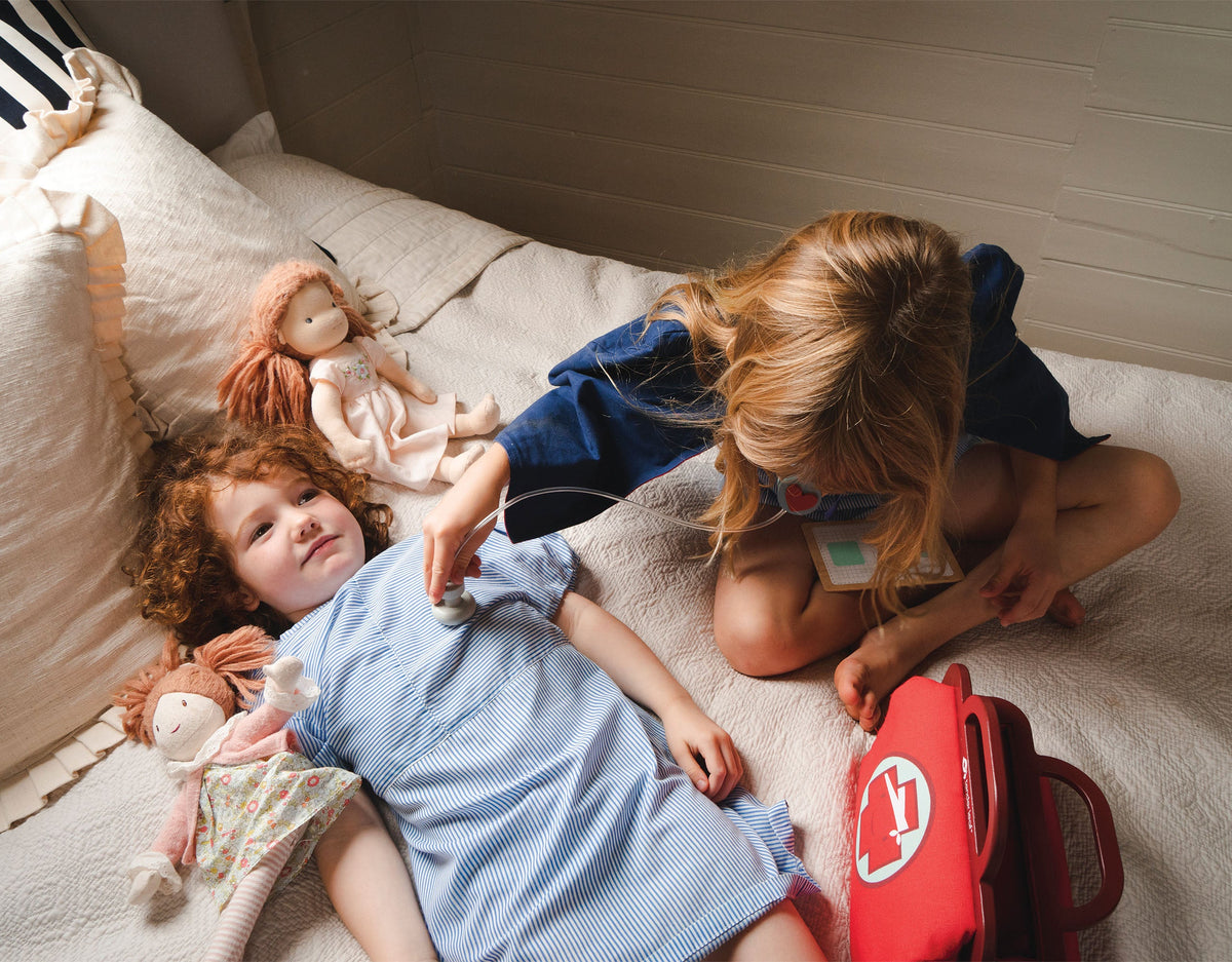 Two children playing with dolls on a bed, one child holding a red toy bag.