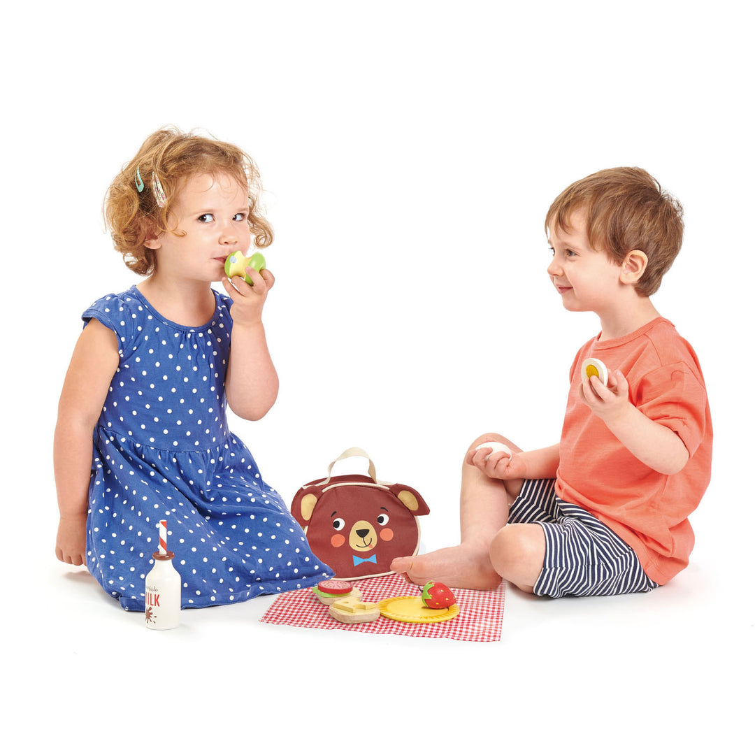Two children sitting on a white background with a teddy bear picnic set.