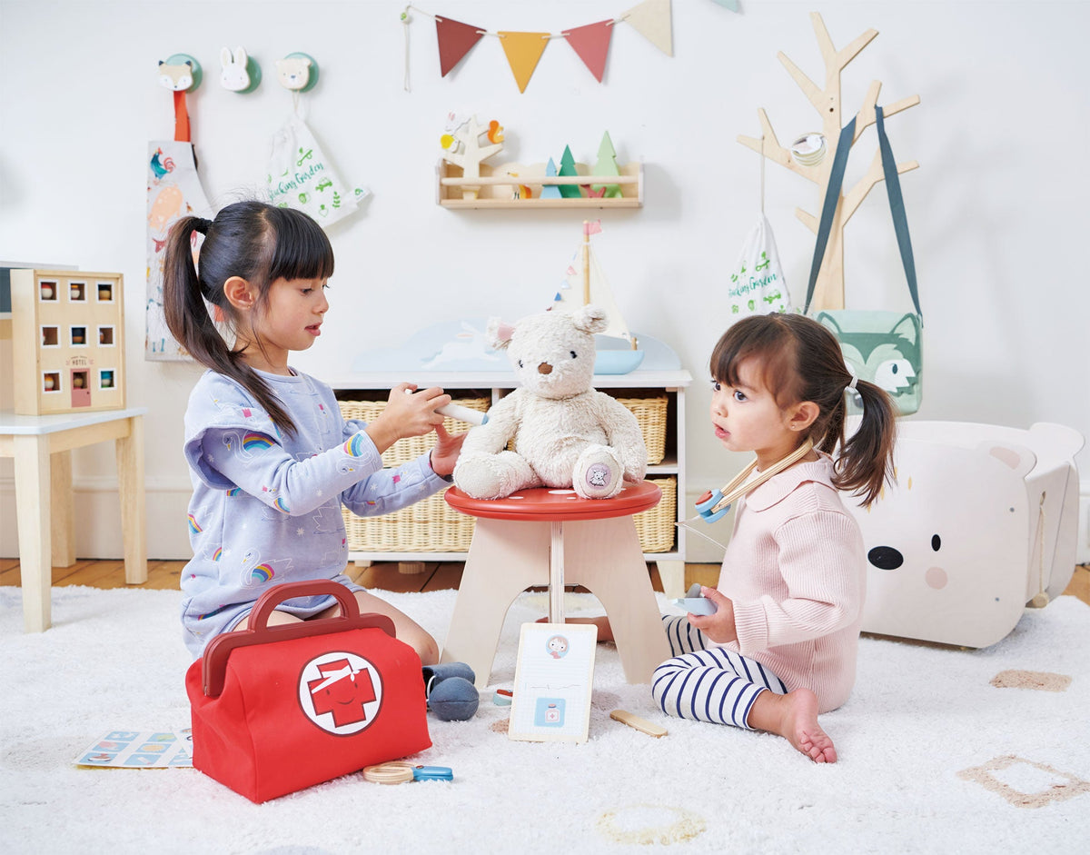 Two children playing with a red toy first aid kit in a room with toys and furniture.