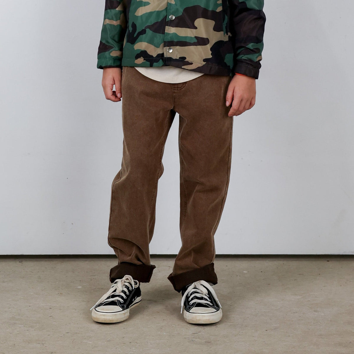 Person wearing a camouflage jacket, brown pants, and black sneakers on a plain background