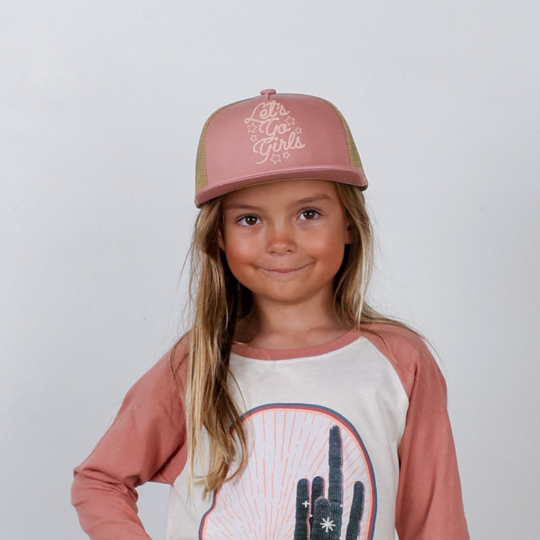 Young girl wearing a pink cap with text and a graphic t-shirt on a plain background