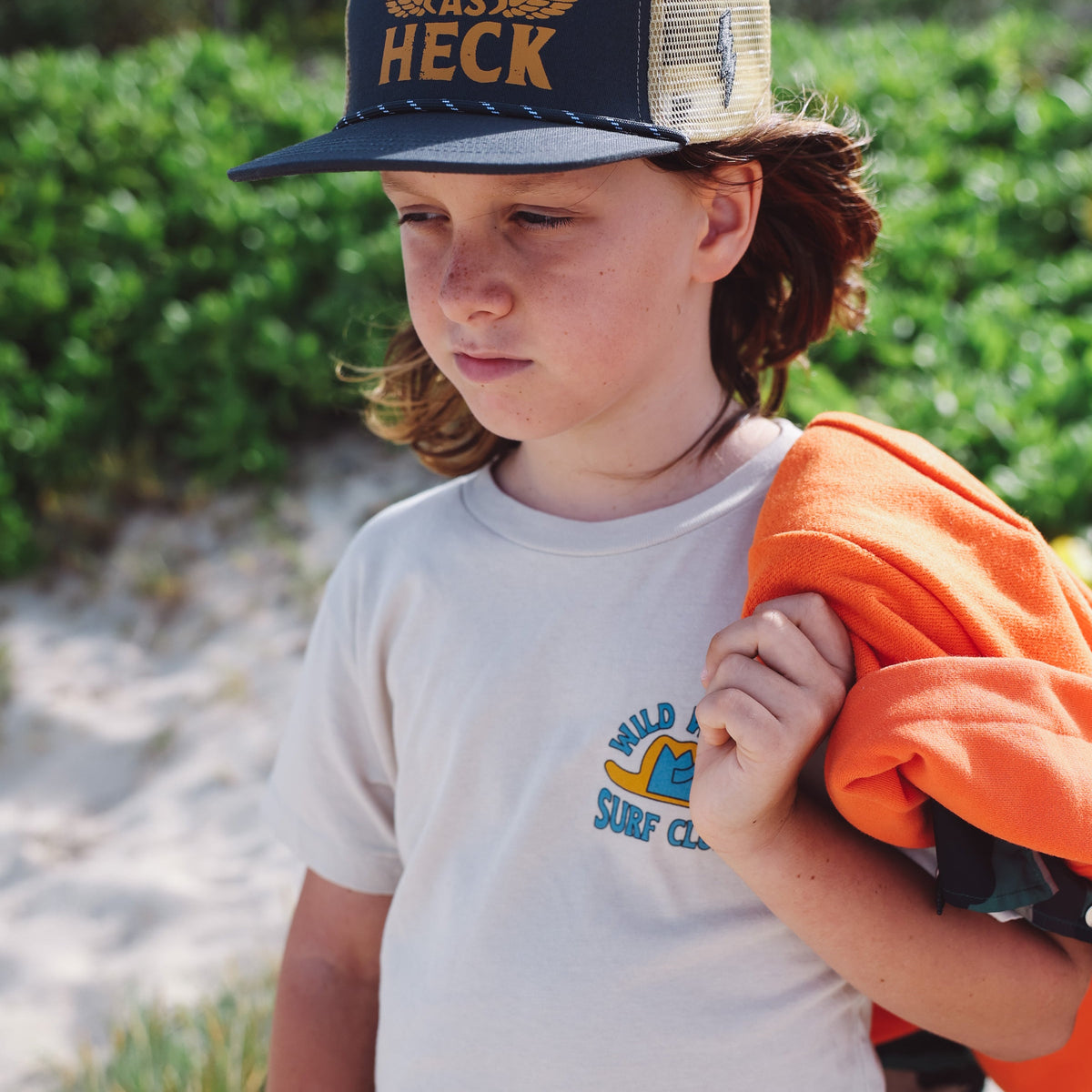 Child wearing a 'Wild as Heck' cap and holding an orange shirt on a beach.