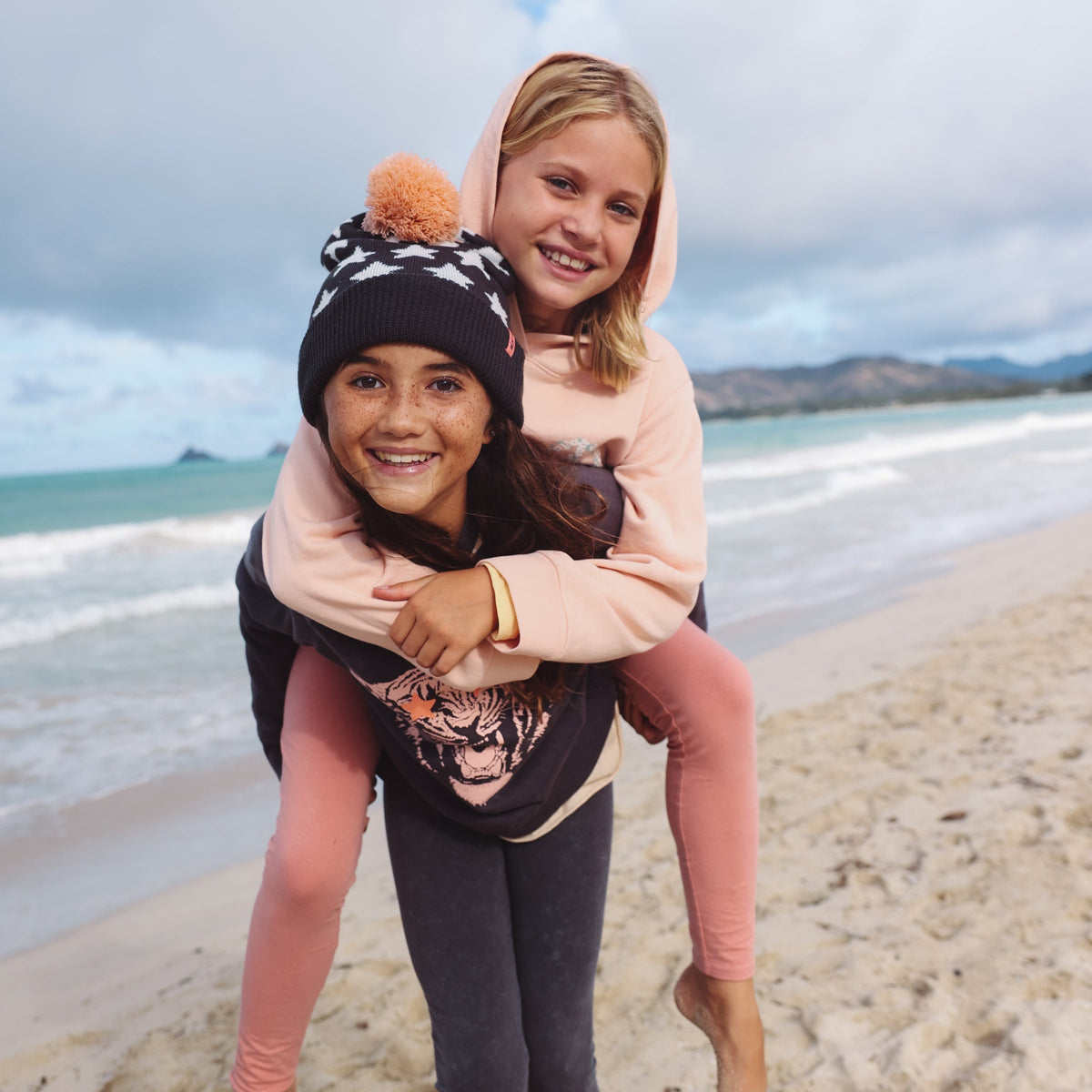 Two young girls on a beach with one girl piggybacking the other, smiling at the camera.