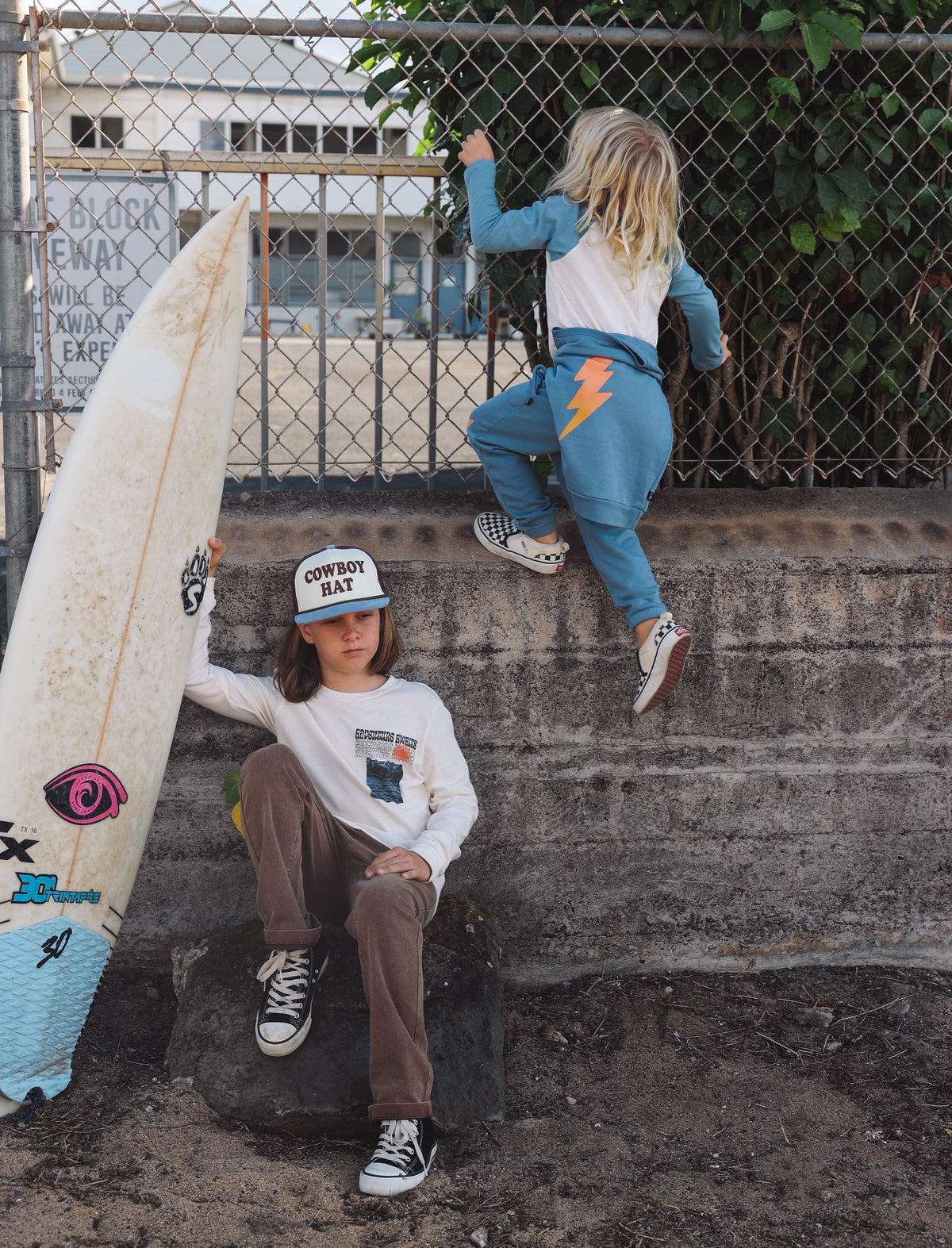 Two children with a surfboard on a concrete surface with a metal fence and building in the background.