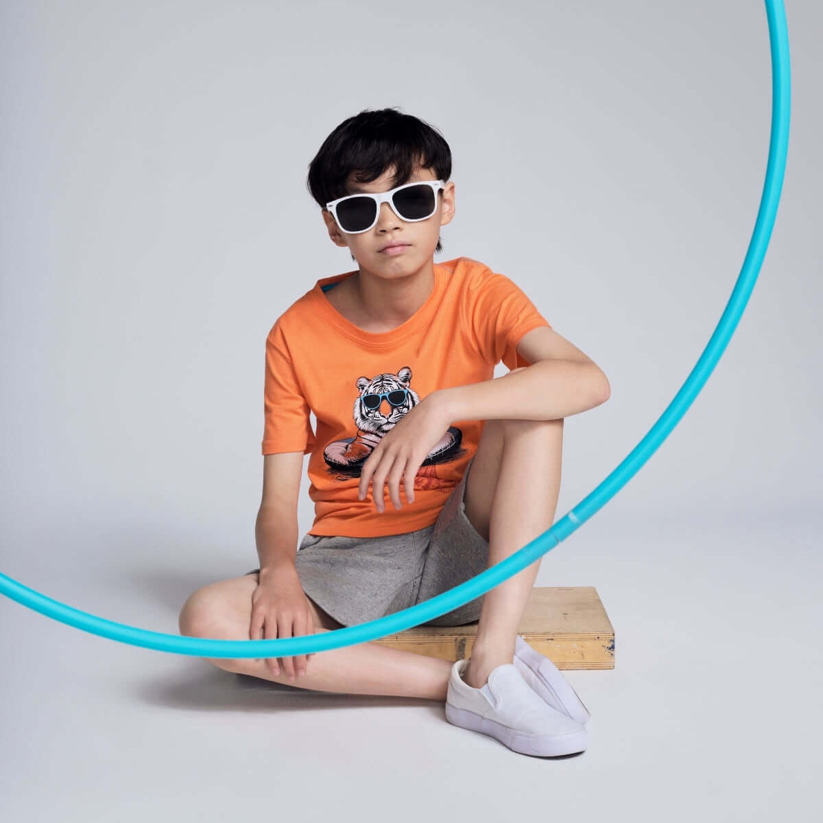 Child wearing an orange t-shirt with a graphic design, sitting on a wooden block with a blue hula hoop.