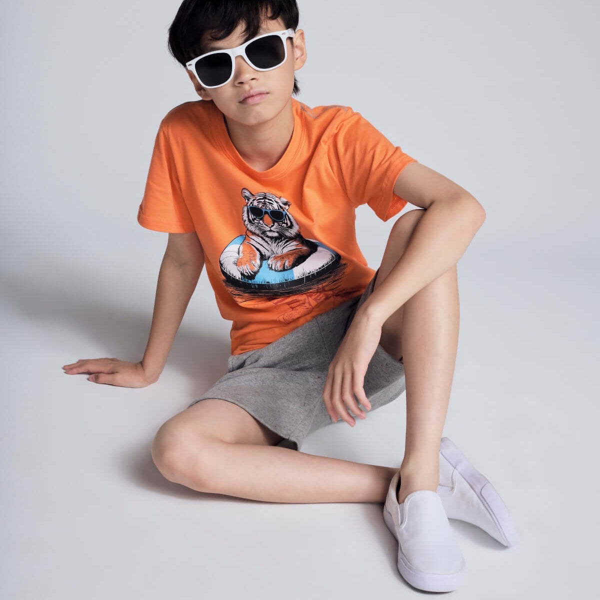 Child wearing an orange t-shirt with a tiger graphic, gray shorts, and white sneakers on a light gray background.
